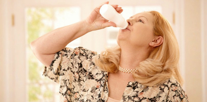 Senior woman standing in kitchen, drinking yogurt in bright background.